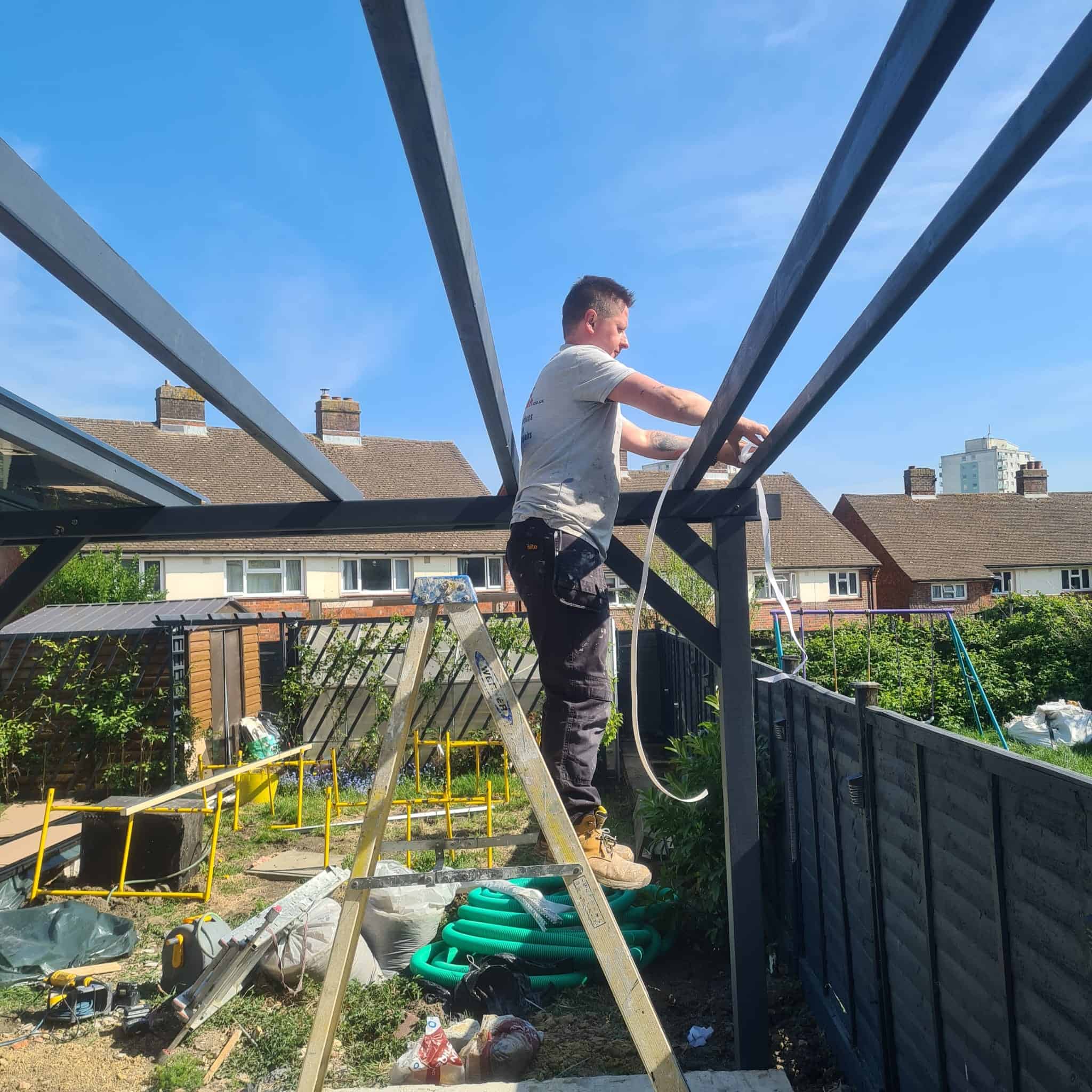 Adam Handyman owner standing on a ladder while working on a steel pergola frame in a residential garden